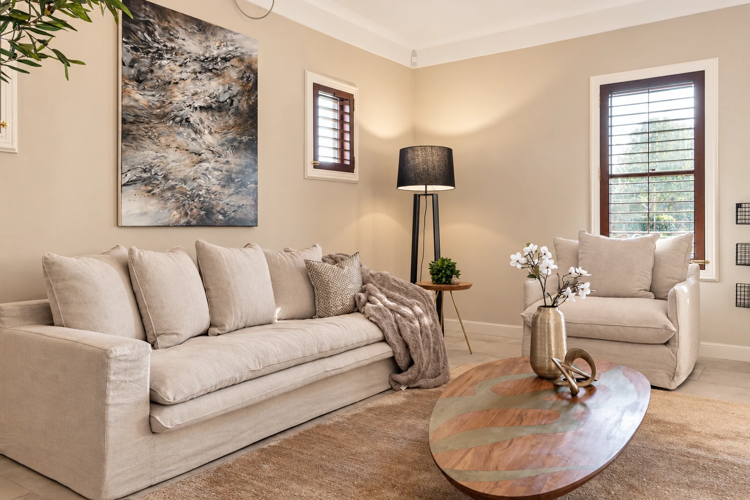 Lounge with a pale linen sofa and armchair, an oval wooden coffee table, abstract artwork and a dark floor lamp.