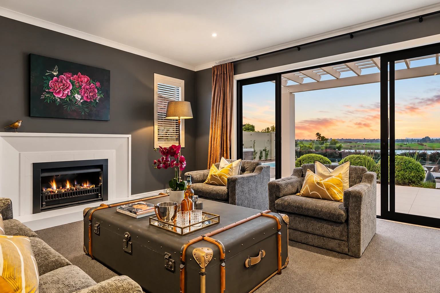 Living room with a charcoal fireplace wall, a trunk coffee table, grey armchairs and yellow cushions beside wide glass doors.