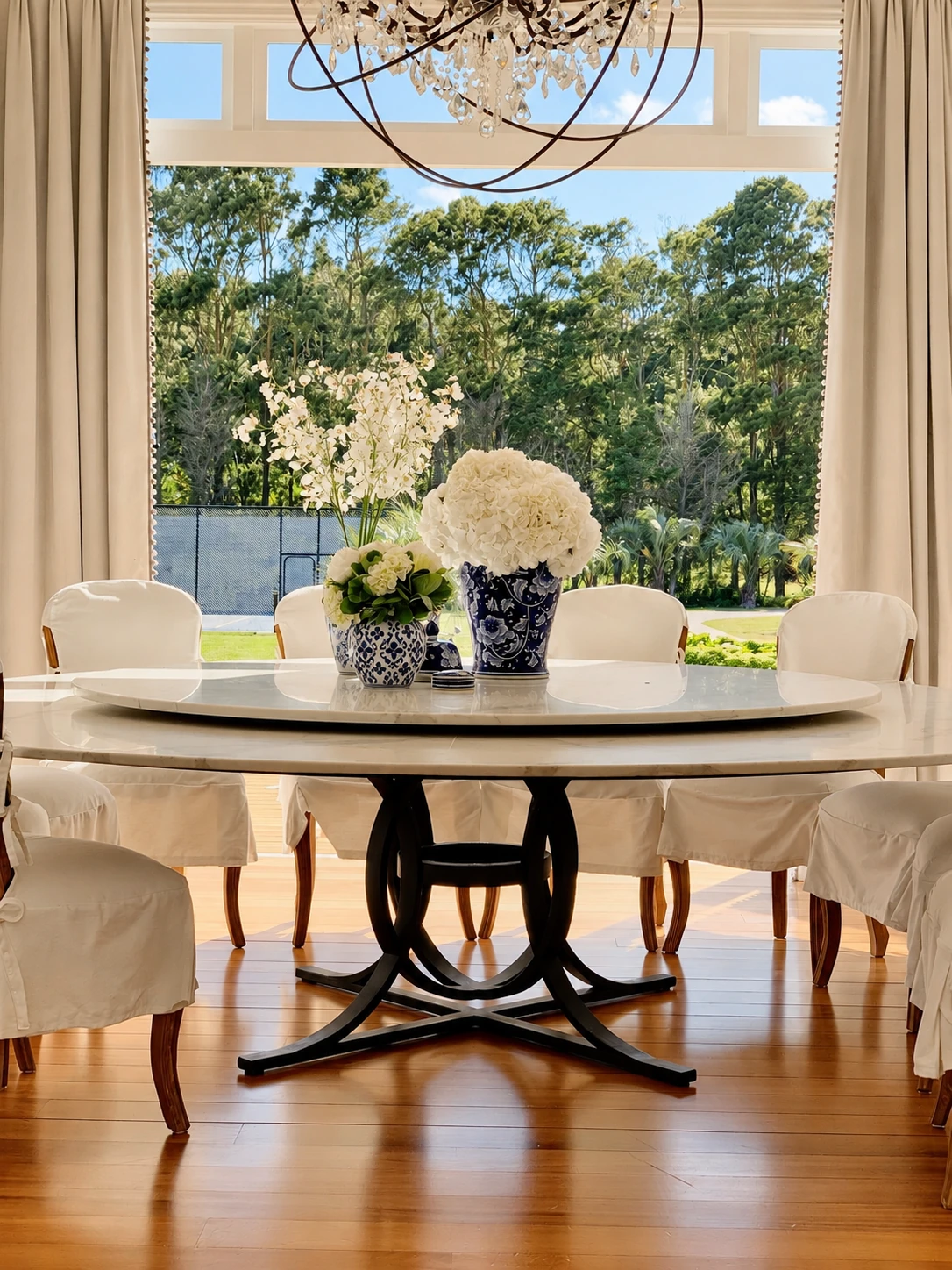 Dining room with a round table, white slipcovered chairs and a crystal chandelier beside tall windows overlooking trees.