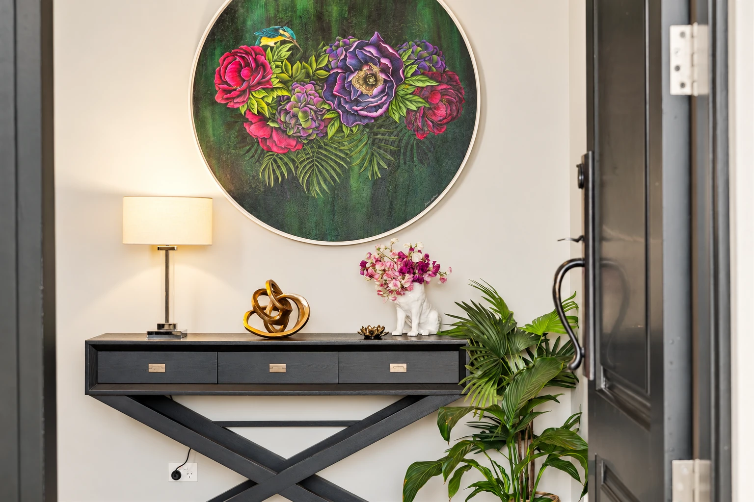 Entrance with a dark console table, circular floral artwork, a table lamp, gold sculpture, flowers and a leafy plant.