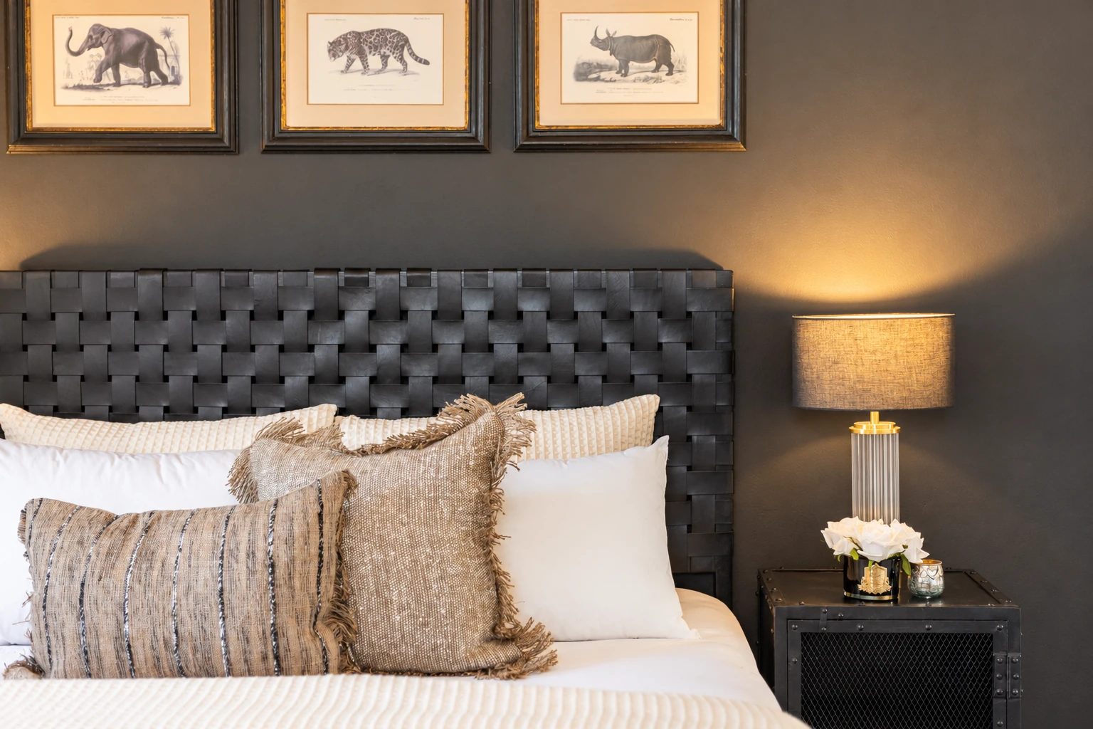 Bedroom details with a dark woven headboard, layered ivory and textured cushions, framed animal prints and a bedside lamp.